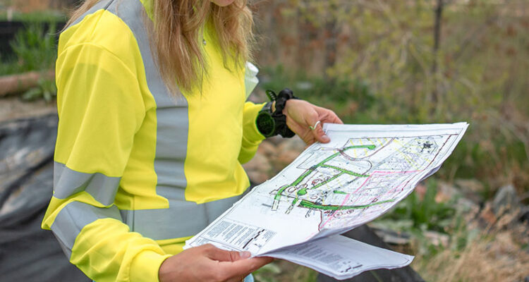 Worker in hard hat and vest studies site map outdoors.