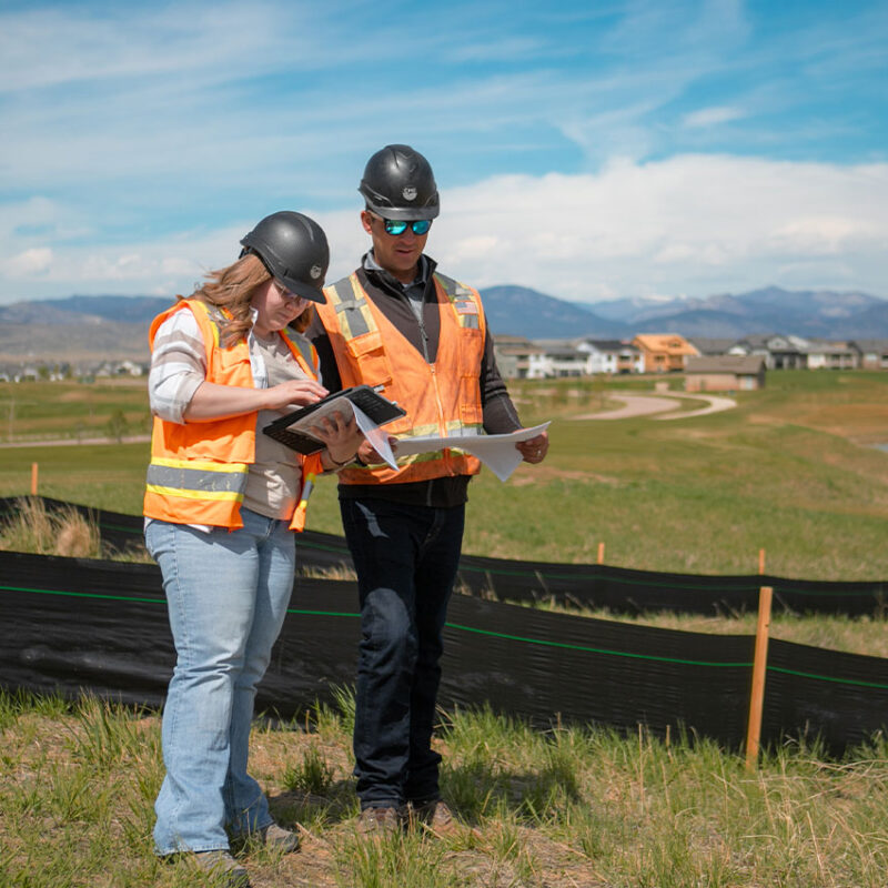 Two construction workers review documents at a grassy outdoor site.