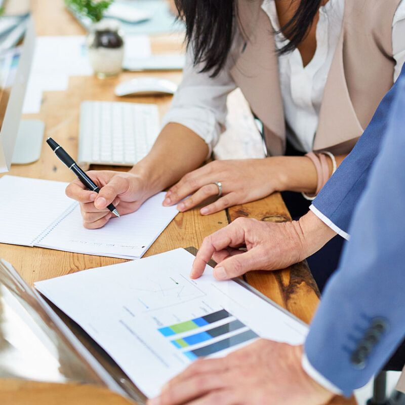 Two reviewing charts at desk; one writes, one points; computer behind.
