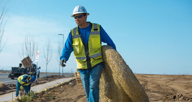Construction worker carries coiled material; another stands in background.