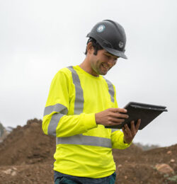 Worker in yellow vest and hard hat uses tablet at site.