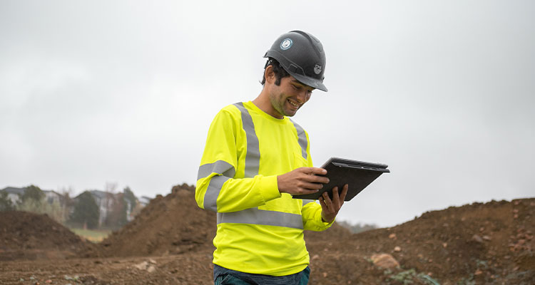 Worker in yellow vest and hard hat uses tablet at site.