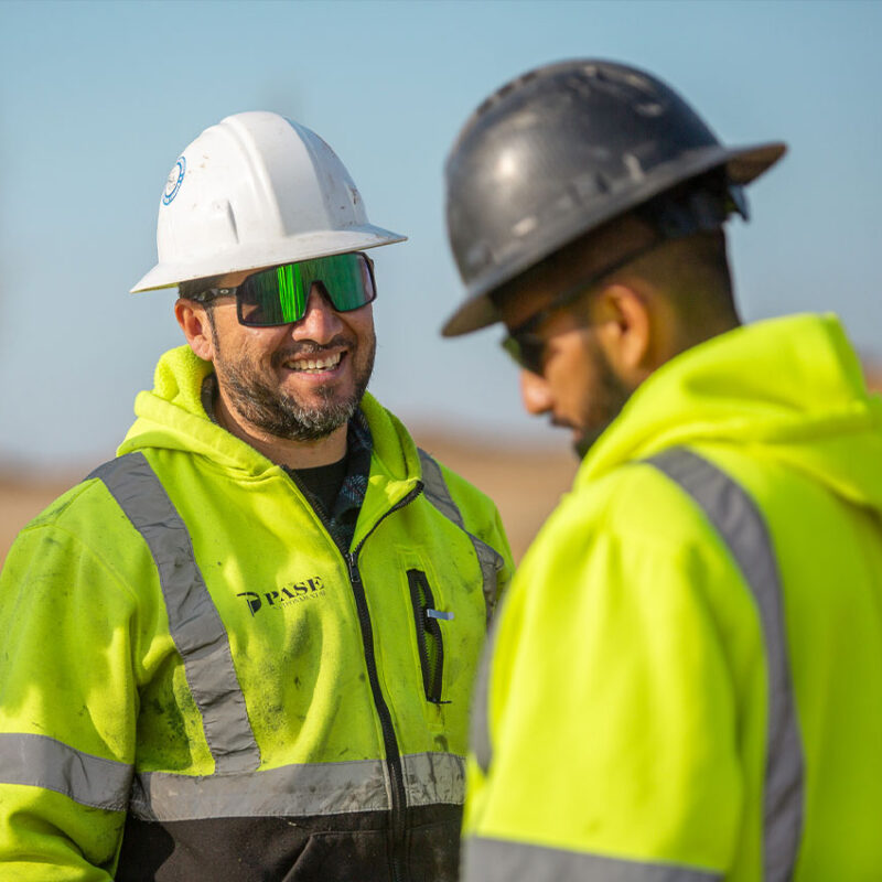 Two workers in hi-vis jackets and hard hats talking outside.