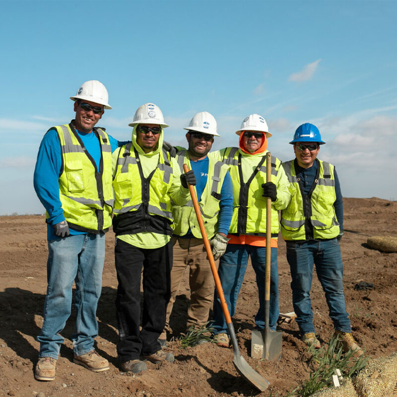 Five construction workers in safety gear pose with shovels.
