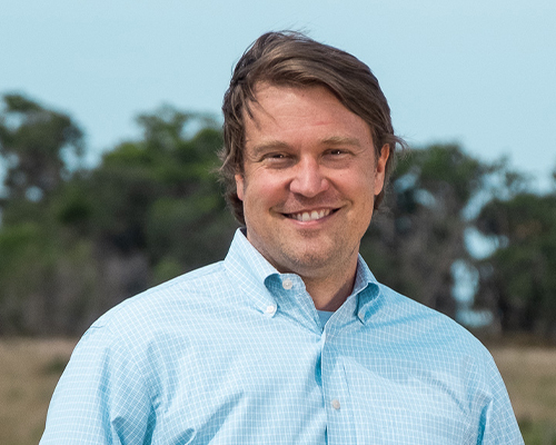 Man in light blue shirt standing outside with trees and sky.