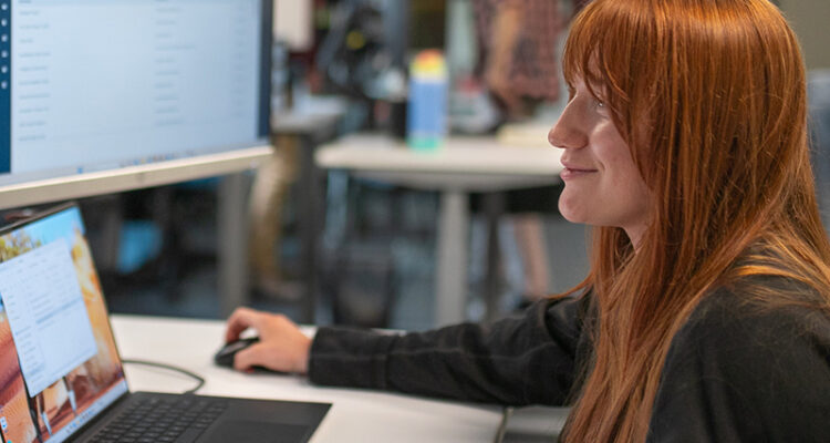 Red-haired woman at desk with laptop; two others in background.