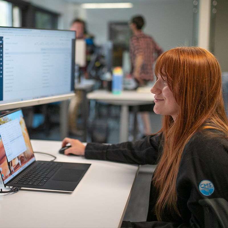 Red-haired woman at desk with laptop; two others in background.
