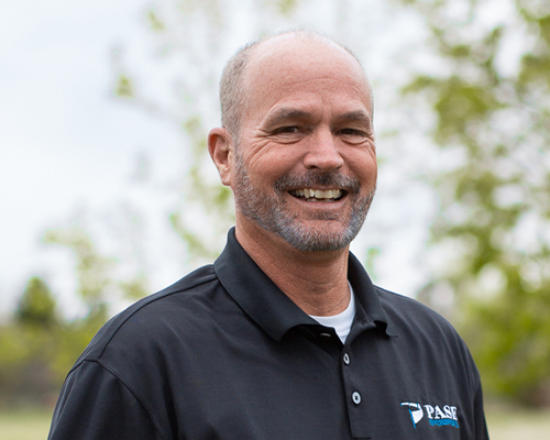 Smiling middle-aged man with gray beard, PASE shirt, outdoors.