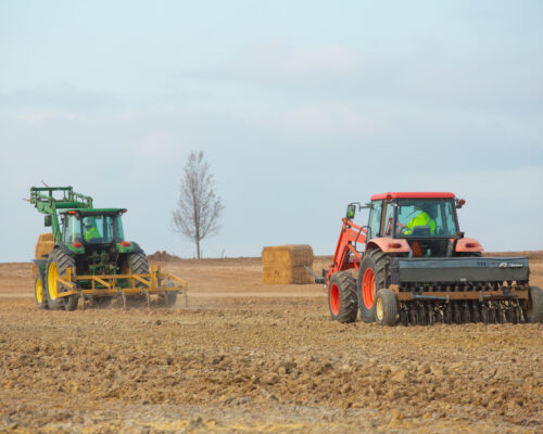 Two tractors in a field; hay bales and tree behind.