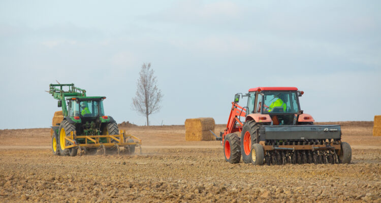 Two tractors in a field; hay bales and tree behind.