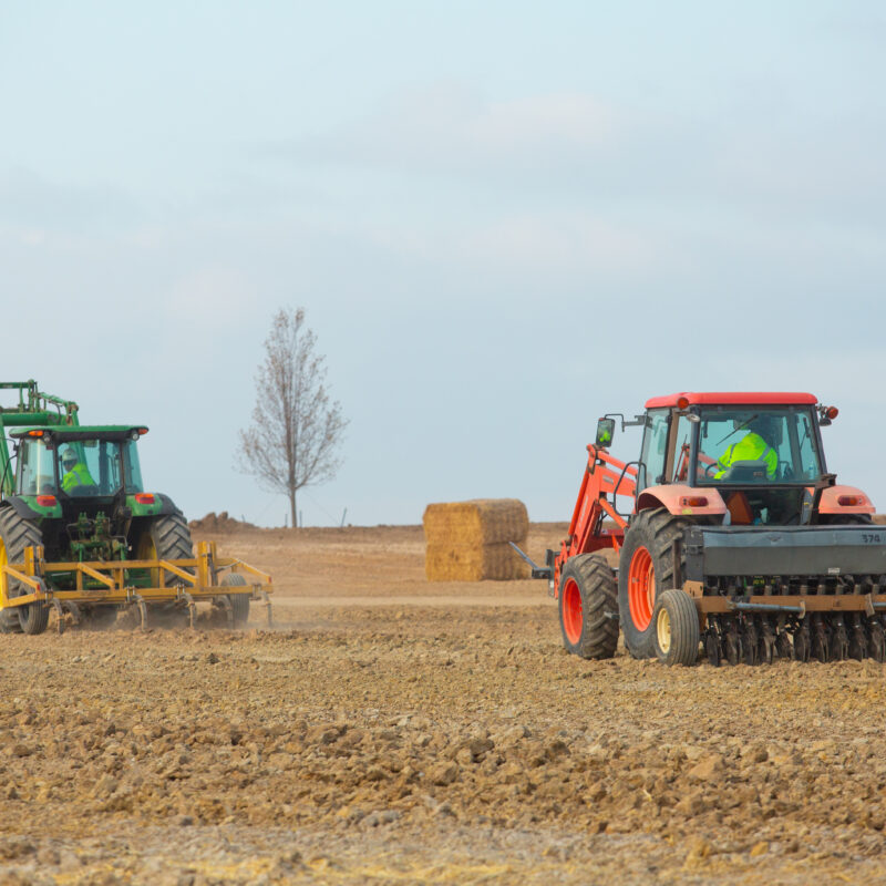 Two tractors in a field; hay bales and tree behind.