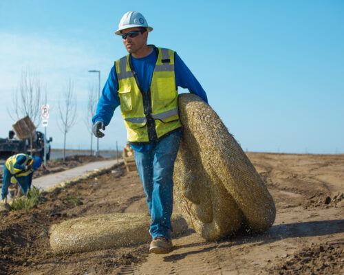 Worker in vest and helmet carries straw rolls on dirt site.