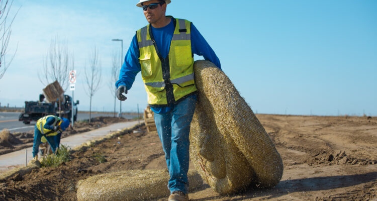 Worker in vest and helmet carries straw rolls on dirt site.